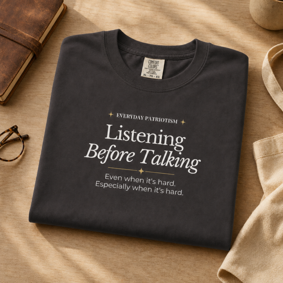 A folded dark charcoal gray graphic T-shirt is styled on a beige fabric surface with a brown leather notebook, eyeglasses, and a beige tote bag nearby. The shirt has white text reading 'Listening Before Talking' with smaller text above and below, and a small neck label is visible.