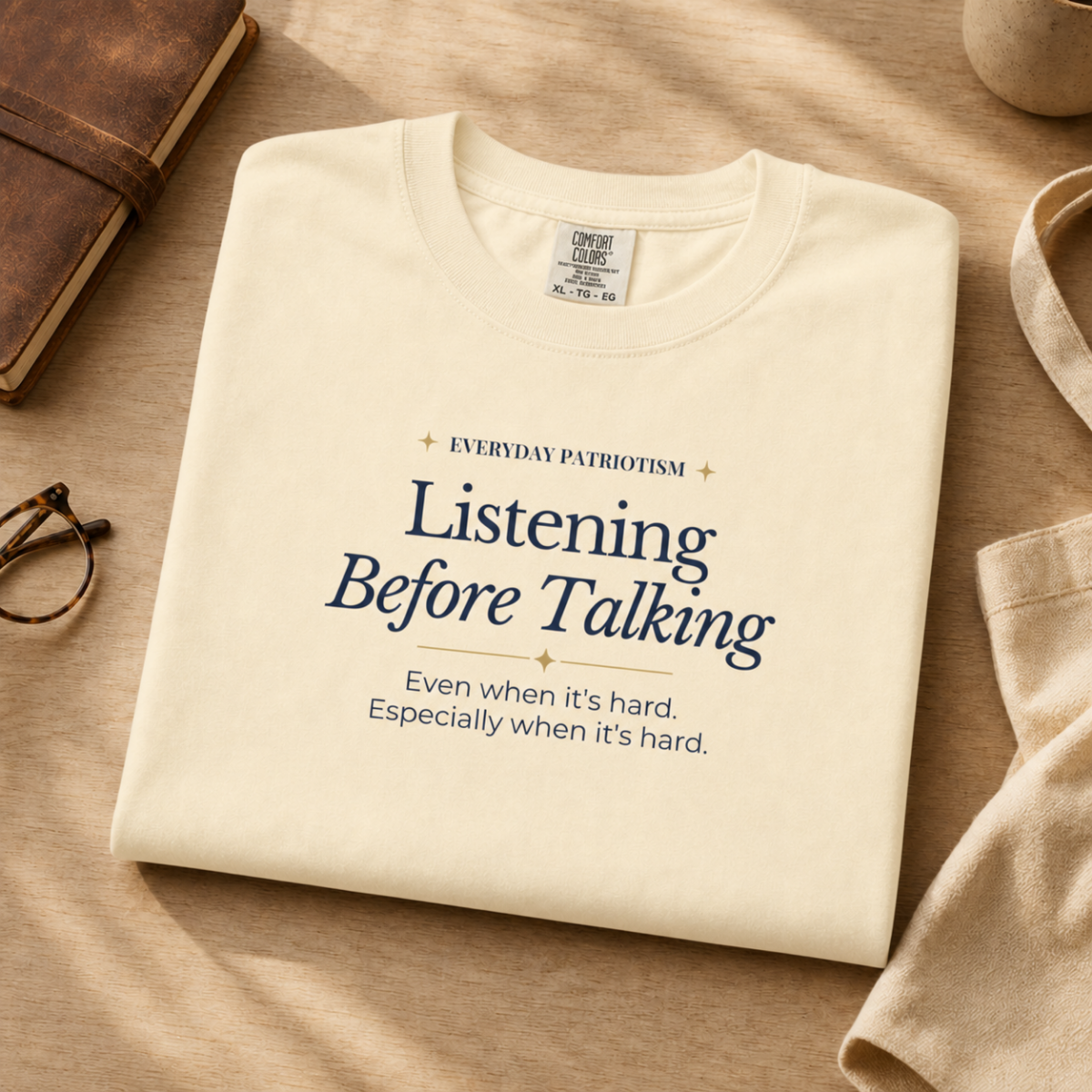 A folded cream-colored t-shirt with navy blue text reading 'Listening Before Talking' and a motivational quote underneath, styled on a beige fabric surface with a brown notebook, eyeglasses, and a beige bag nearby.