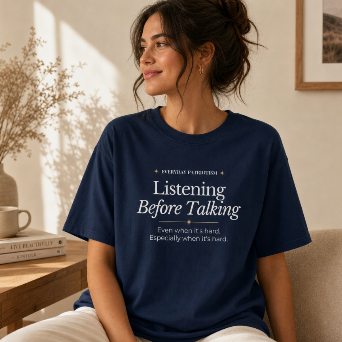 A woman sits indoors wearing a navy blue graphic T-shirt with the phrase 'Listening Before Talking' printed in white text on the front. The setting is a warm, sunlit living space with a table, books, dried flowers, and a framed picture in the background.
