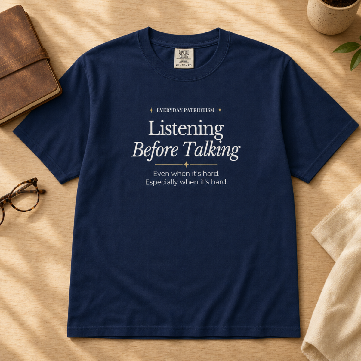 A navy blue graphic T-shirt laid flat on a beige fabric surface with styled props including a brown notebook, eyeglasses, a small plant, and a folded cream cloth. The shirt has white printed text reading 'Listening Before Talking' with smaller lines above and below.