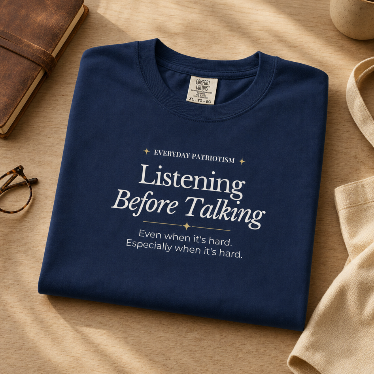 A navy blue folded T-shirt with white printed text reading 'Listening Before Talking' and smaller slogan text below, styled on a warm wood surface with a notebook, eyeglasses, and fabric props nearby.