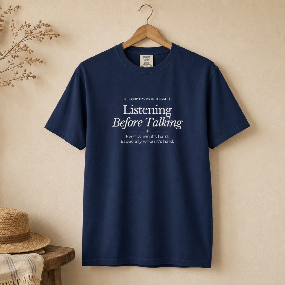 A navy blue short-sleeve t-shirt hanging on a wooden hanger against a warm beige wall. The shirt features white printed text on the chest that reads 'LISTENING Before Talking Even when it's hard.