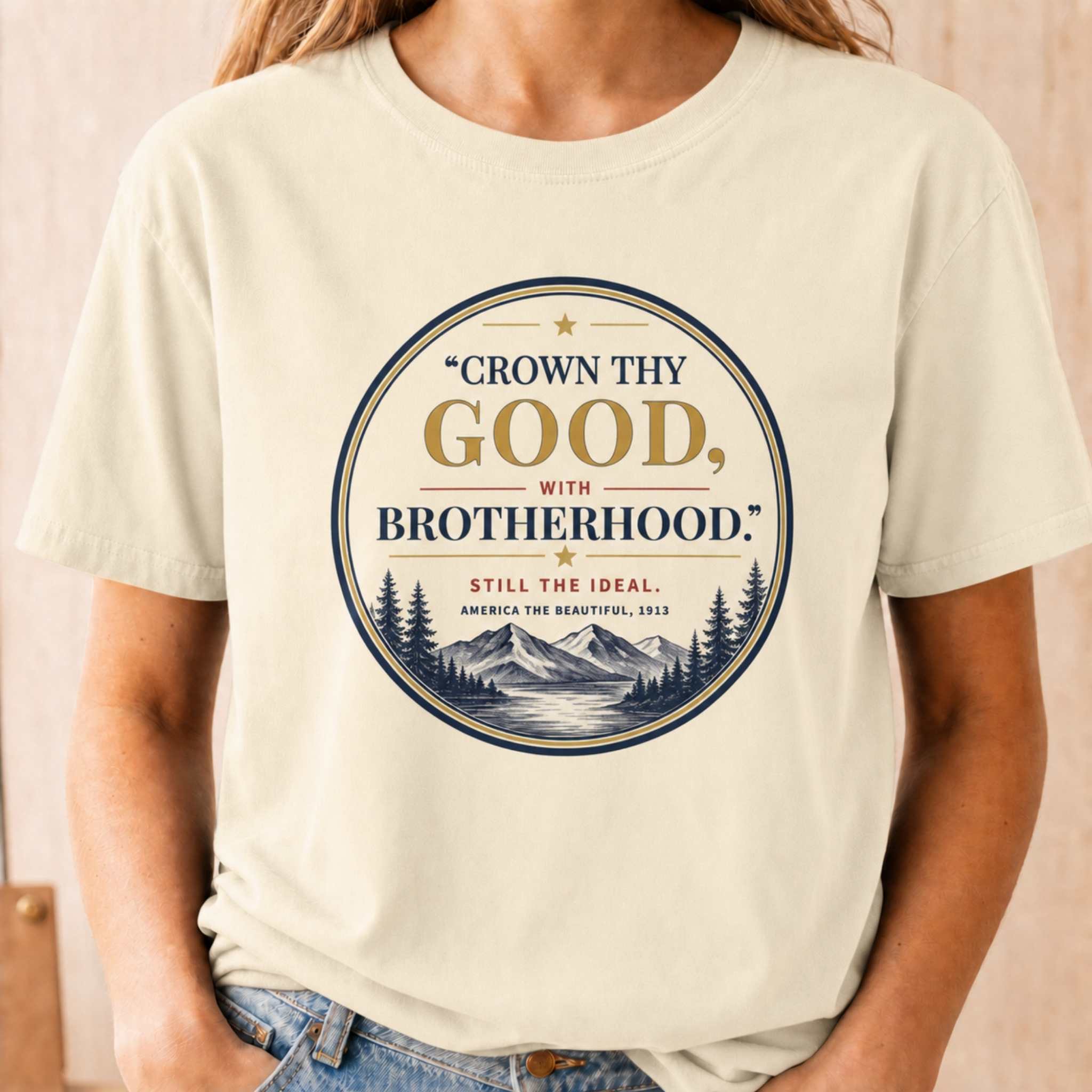 A person wearing a cream-colored short-sleeve t-shirt with a circular graphic print that reads "CROWN THY GOOD, WITH BROTHERHOOD." and features a mountain and pine tree illustration.