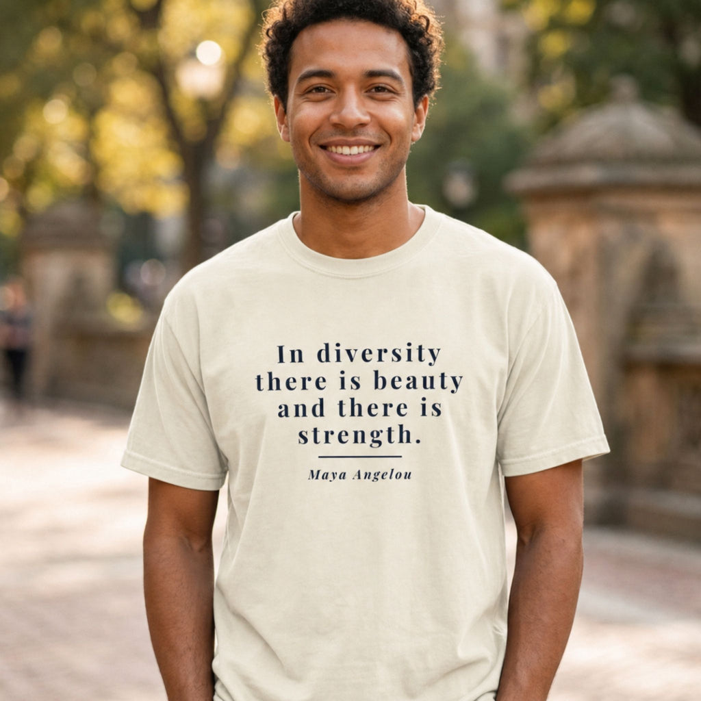 A smiling man wearing a cream-colored t-shirt with a dark navy blue quote from Maya Angelou "In diversity there is beauty and there is strength" printed on the front, set against a blurred outdoor background of trees and a stone structure.