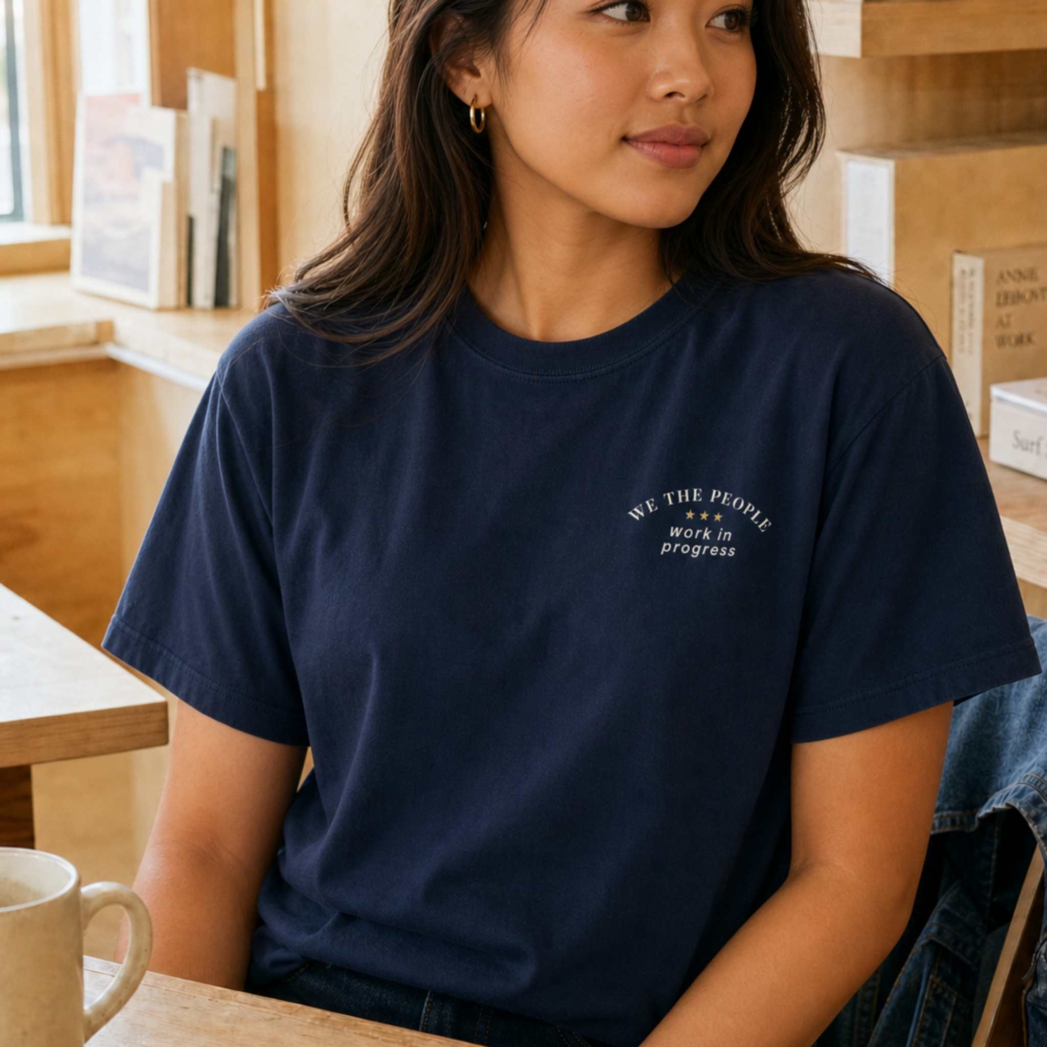 A woman wearing a navy blue crewneck t-shirt with a small white graphic on the left chest that reads 'WE THE PEOPLE WORK IN PROGRESS' with three stars, sitting in a warm, sunlit cafe environment.